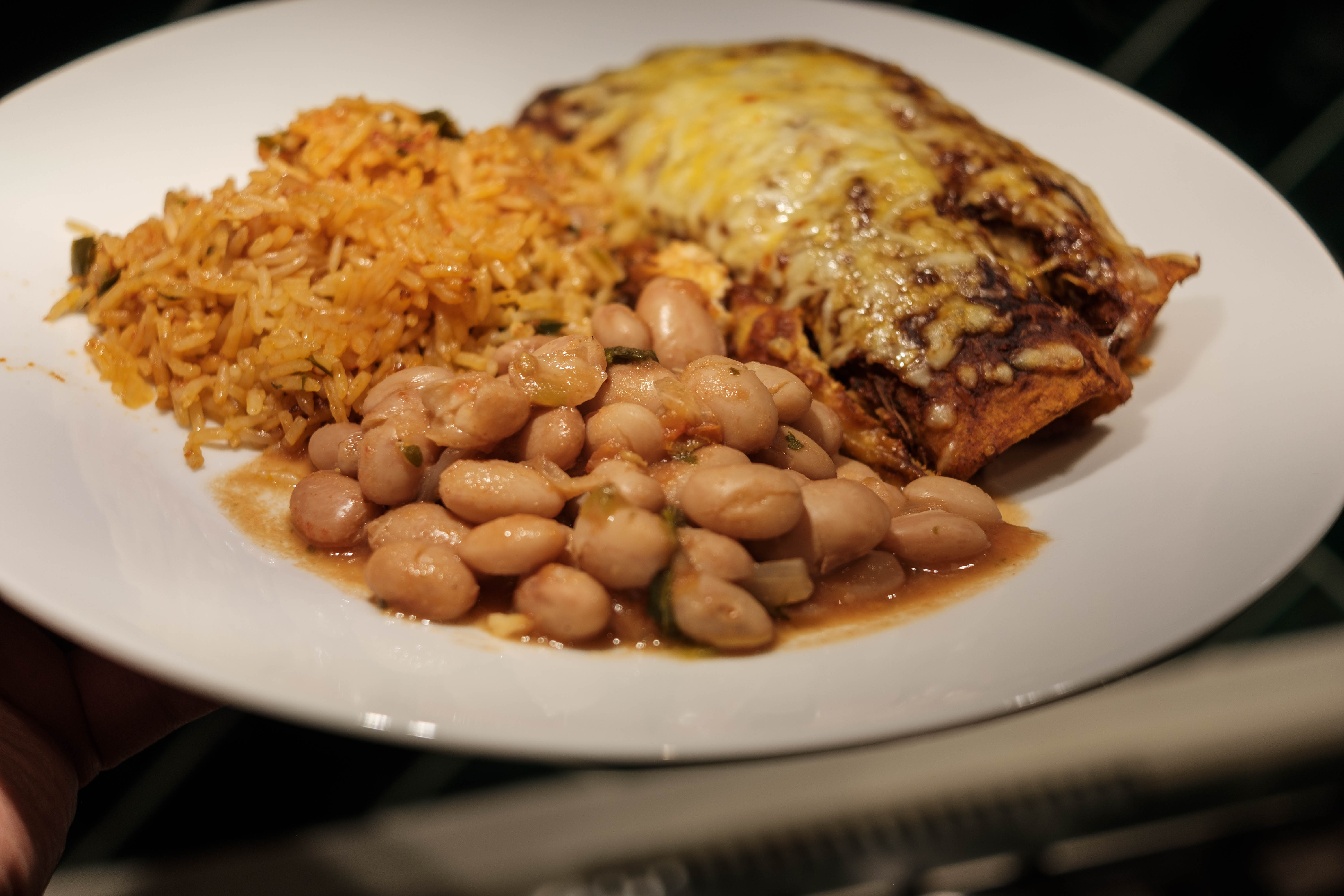 A close-up image of a plated meal, showing a generous portion of sous vide pinto beans alongside golden-brown Mexican rice and a cheese-covered enchilada. The pinto beans are plump and appear tender, nestled in a savory sauce. The enchilada is smothered in melted cheese and browned to perfection, and the rice is speckled with flecks of green herbs. The dish is served on a white plate set against a dark-toned background, creating an inviting contrast that emphasizes the vibrant colors of this TexMex meal.