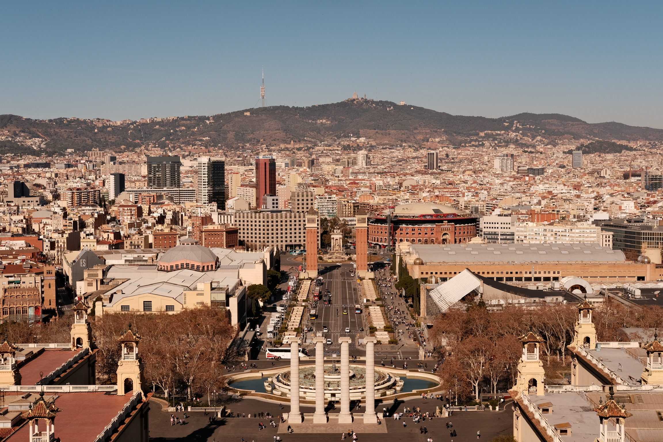 The photo captures a sweeping view of Barcelona, Spain. Dominating the foreground is the Plaça de Catalunya, with its iconic fountain at the center. Surrounding this central square are various city buildings, including the Casa del Rey and the Palau de la Música Catalana. The scene is a lively mix of architecture and urban life. In the distance, the Barcelona Cathedral stands out against the sky, symbolizing the historic charm of the city. The image beautifully encapsulates the essence of this vibrant Spanish city.