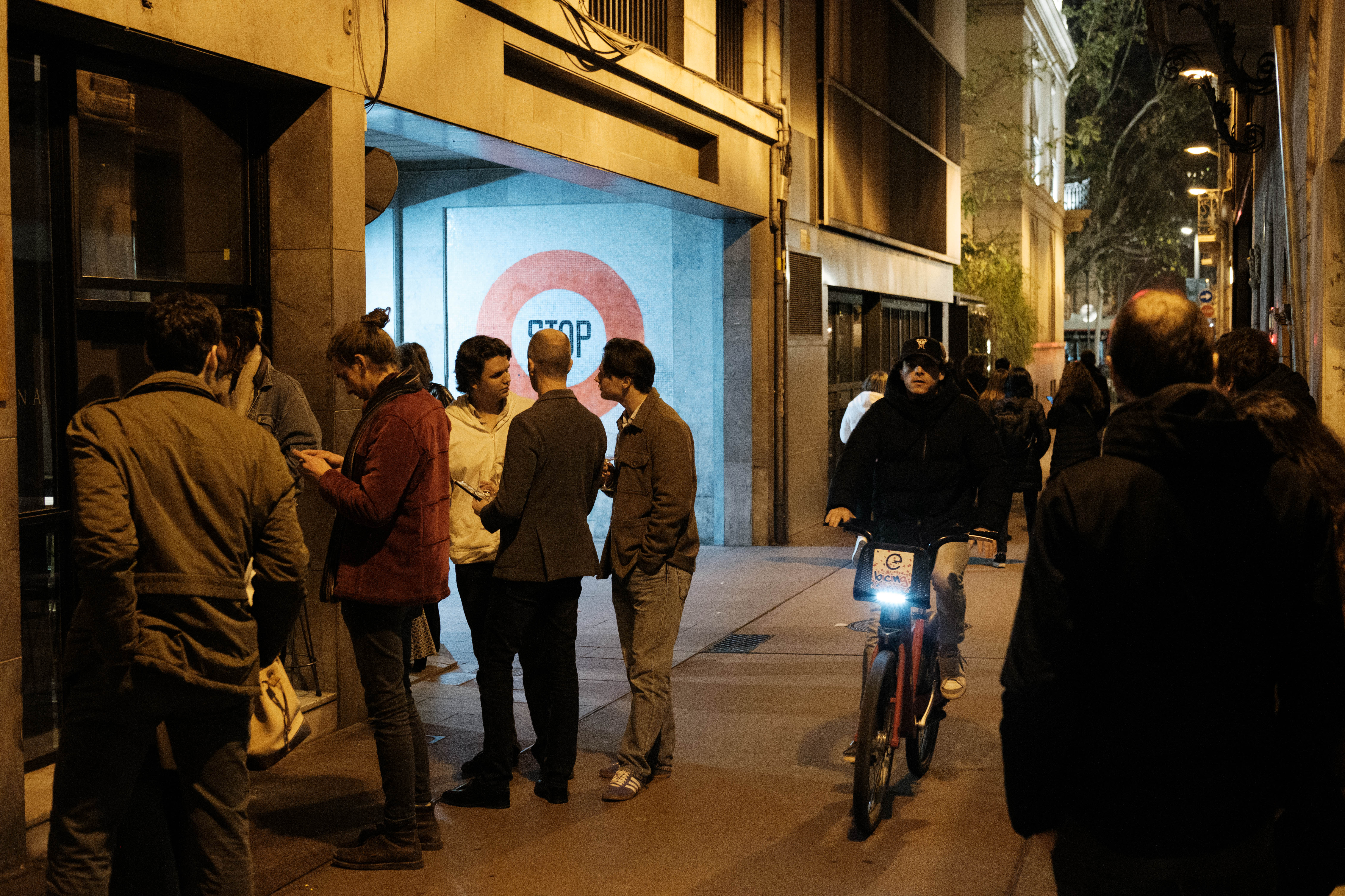 A nighttime scene on a busy street with people standing and conversing. In the foreground, there are individuals engaged in a discussion, with one person holding what appears to be a cell phone. In the background, a group of people is seen near a metro entrance sign. The atmosphere suggests a casual gathering or meet-up, possibly during an event. There are several pedestrians on the street and cyclists passing by. A brightly lit building with a modern design can be observed on the right side of the image, contributing to the urban night setting.