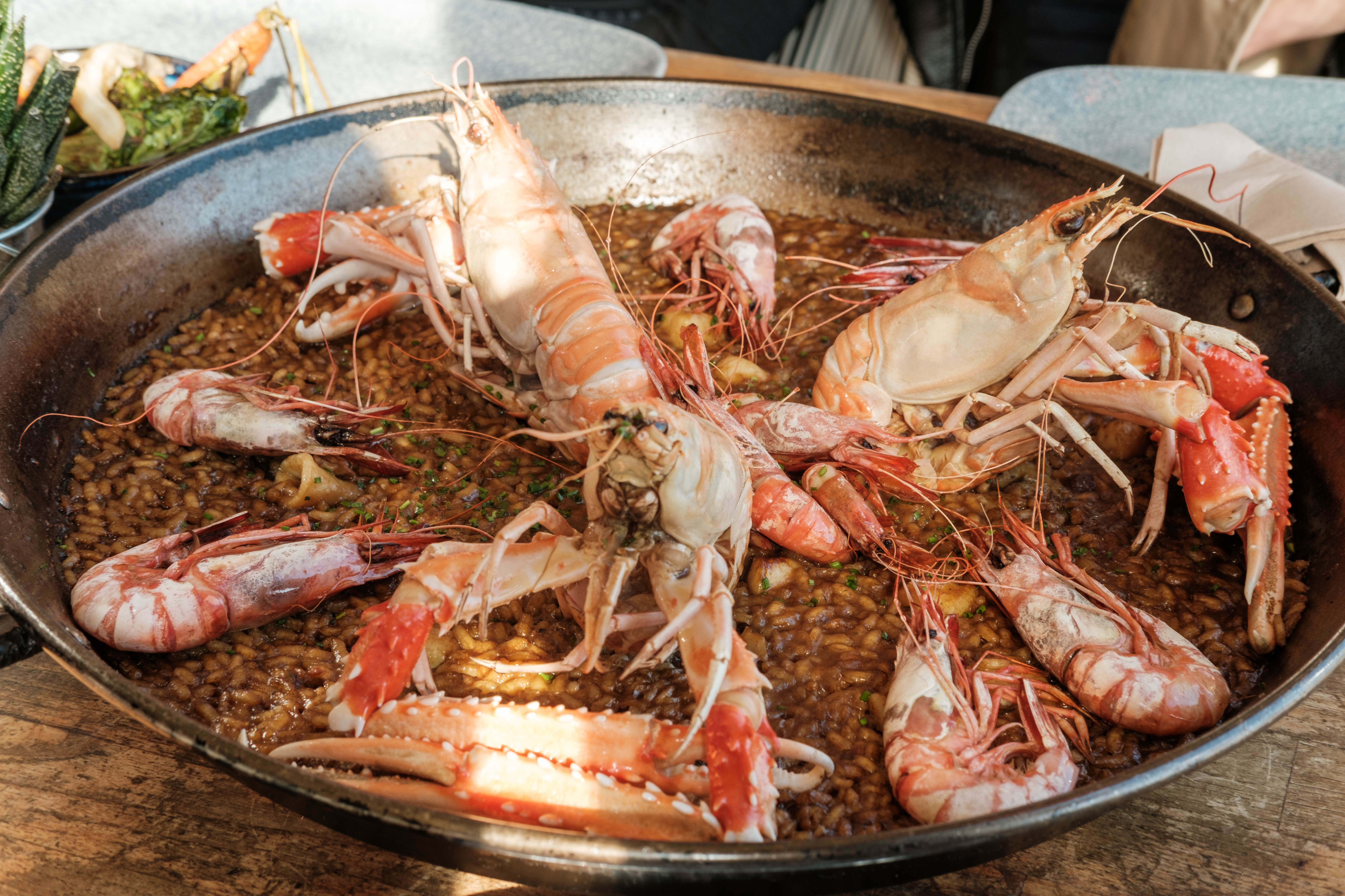 A vibrant dish of paella, a Spanish rice dish, featuring succulent seafood including prawns and crayfish, served on a large metal tray. The rice is speckled with saffron-infused grains and garnished with herbs, creating an appetizing centerpiece for the dining table in the background.