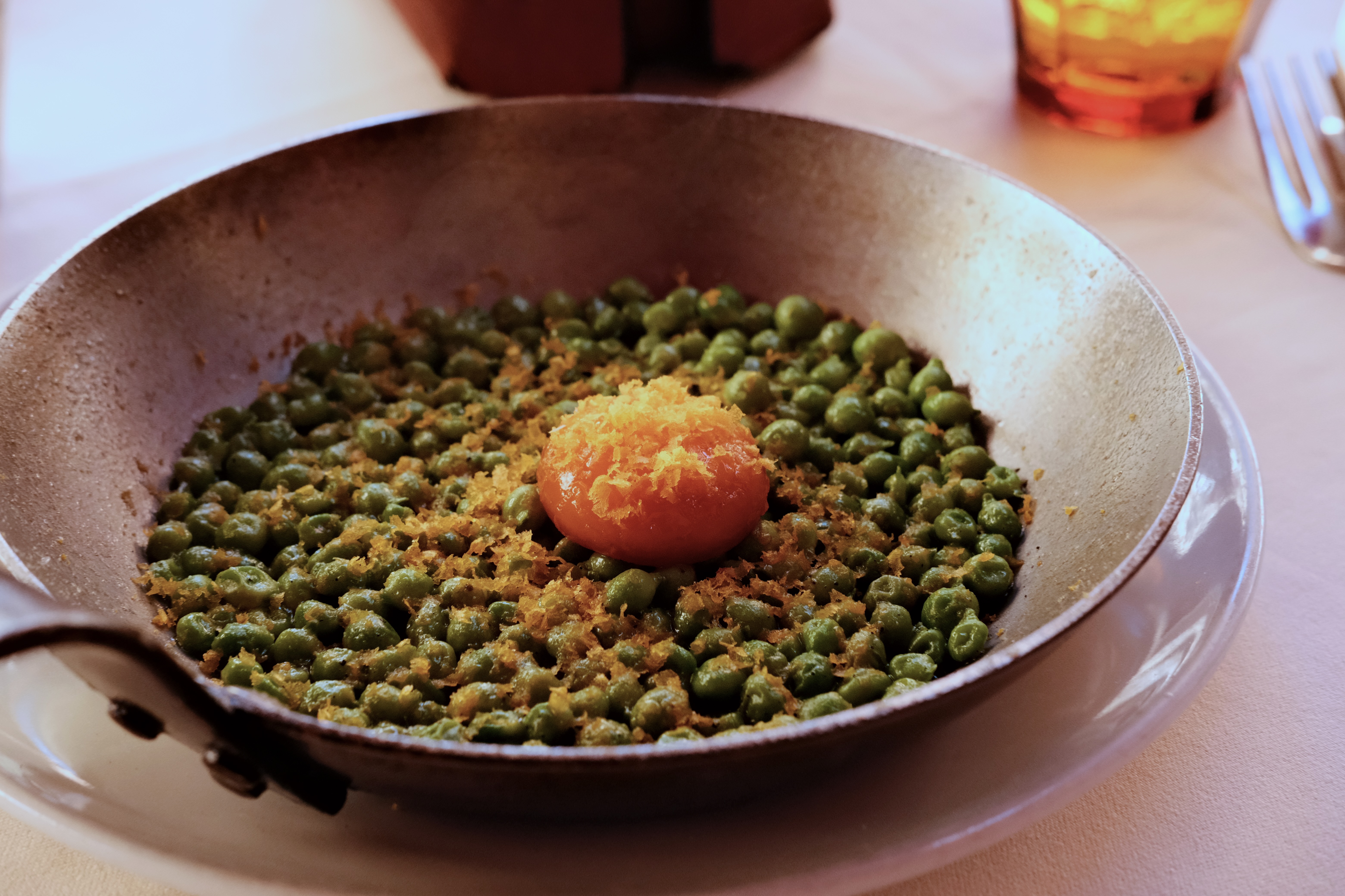 A pan containing fresh peas with an egg yolk on top sprinkled with bottarga set on a dining table in a restaurant.