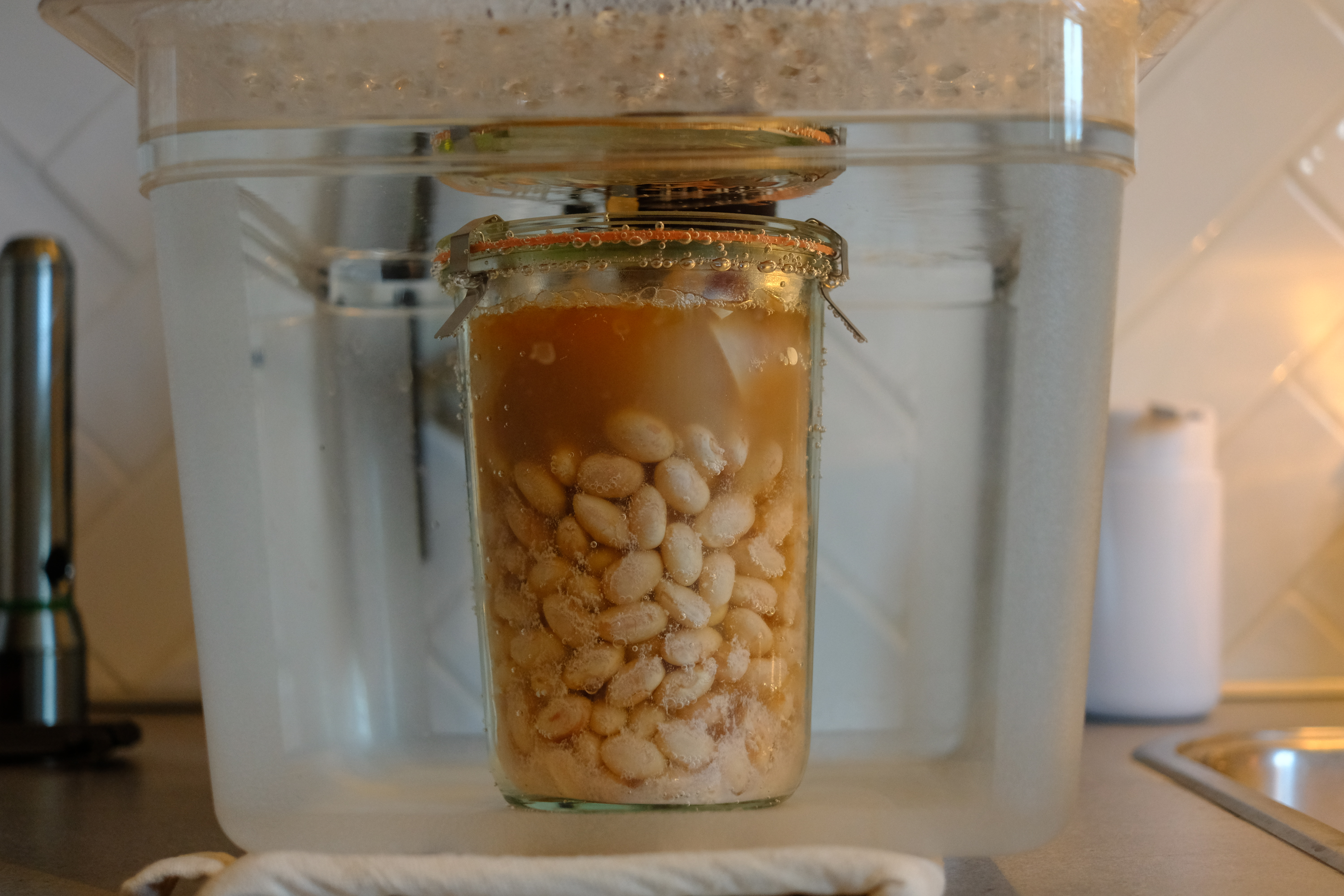 An image showing a jar of pinto beans submerged in a sous vide water bath. The beans appear tender and well-cooked, visible through the clear glass jar, which is sealed with a metal clamp-down lid. Bubbles cling to the sides of the jar, indicating the cooking process. The water bath container is placed on a kitchen countertop, with a white towel underneath it and kitchen tiles in the background.