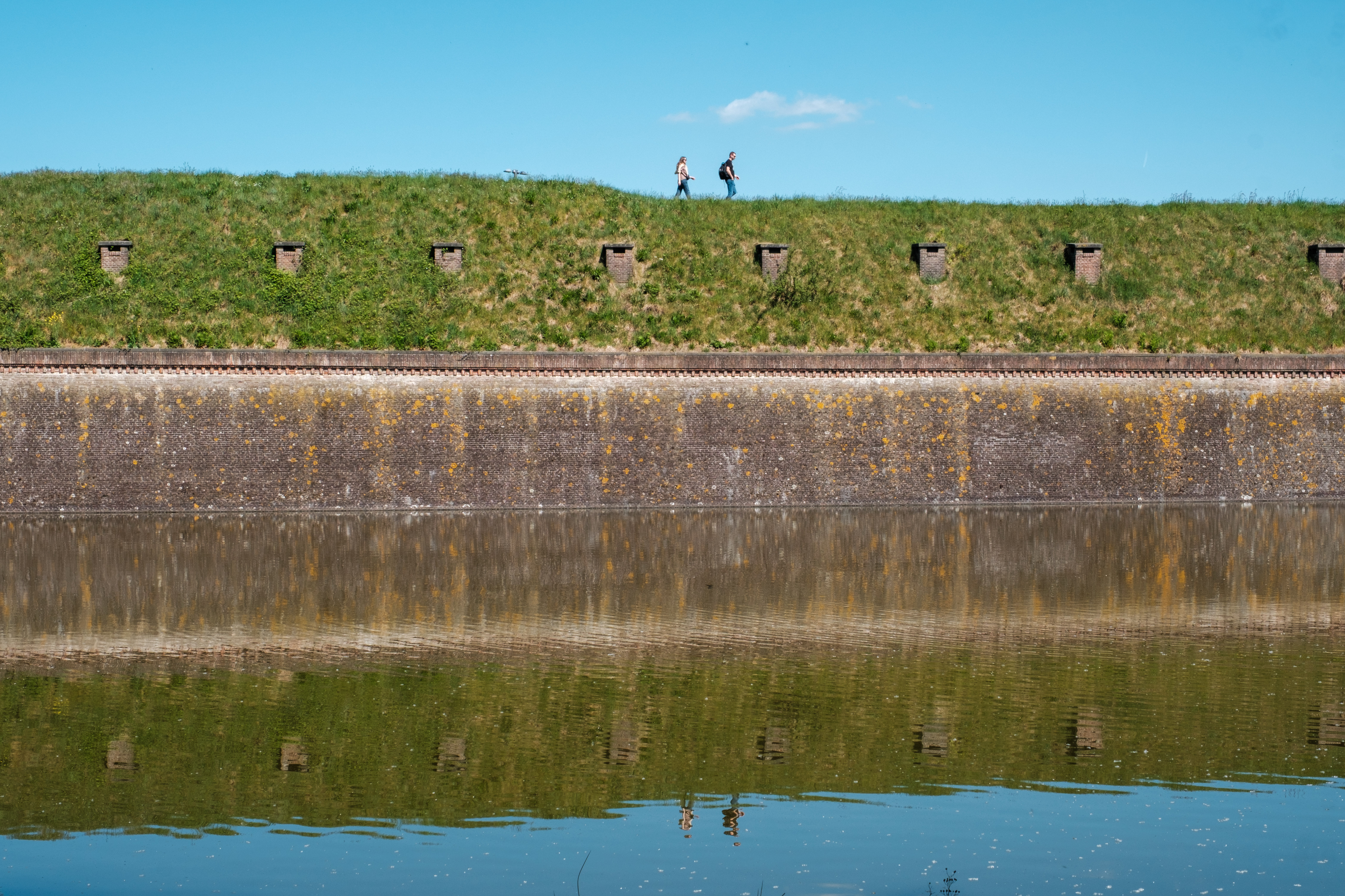 Two people walking along a grassy fortification in Naarden, Netherlands, with a moat reflecting the scene.