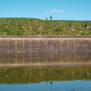 Two people walking along a grassy fortification in Naarden, Netherlands, with a moat reflecting the scene.