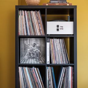 A black shelf with vinyl records, books, a white device, and baskets against a yellow wall.