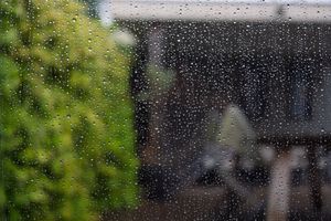 Raindrops on a window with blurred greenery and buildings in the background.